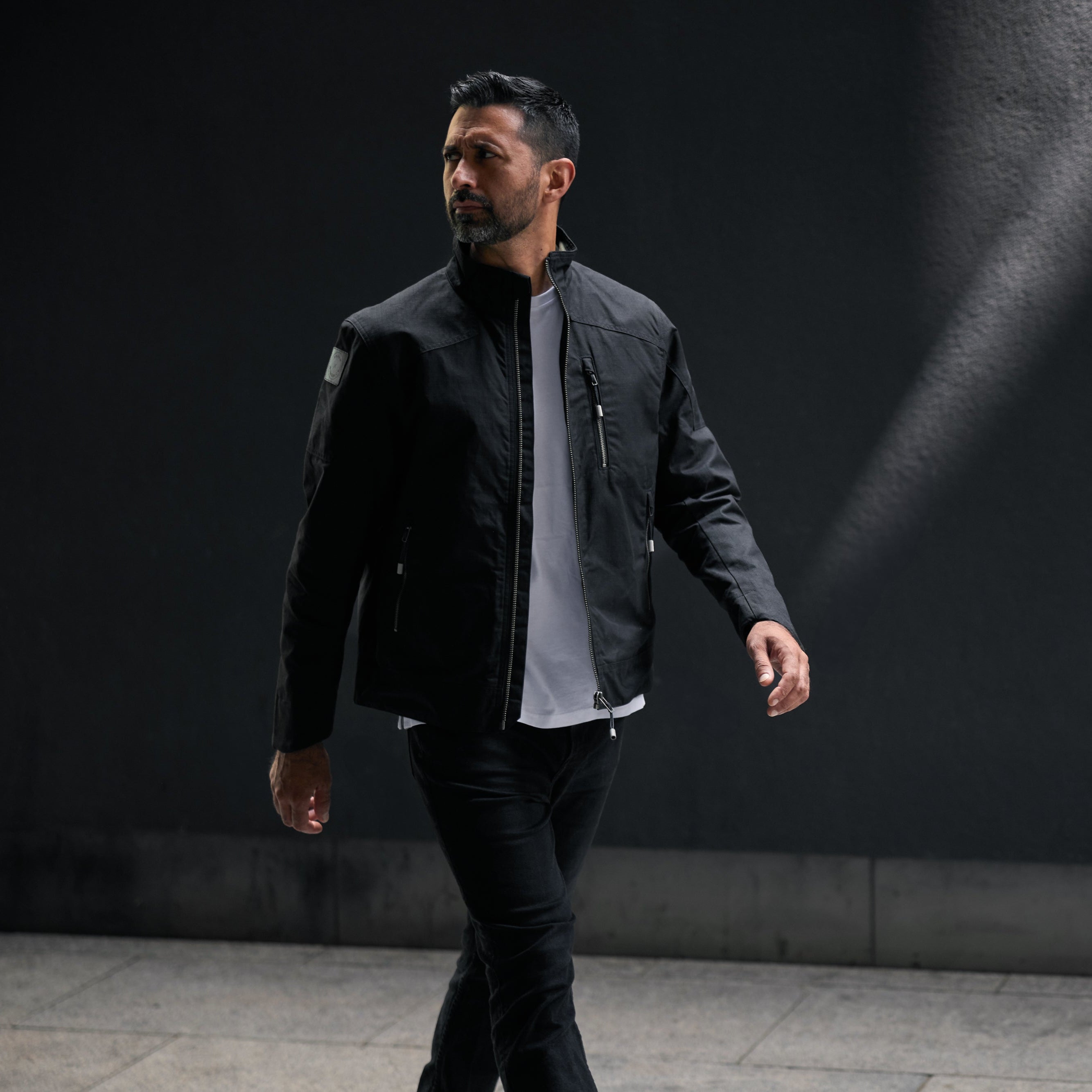 Man wearing a black jacket and dark pants walking against a dark background