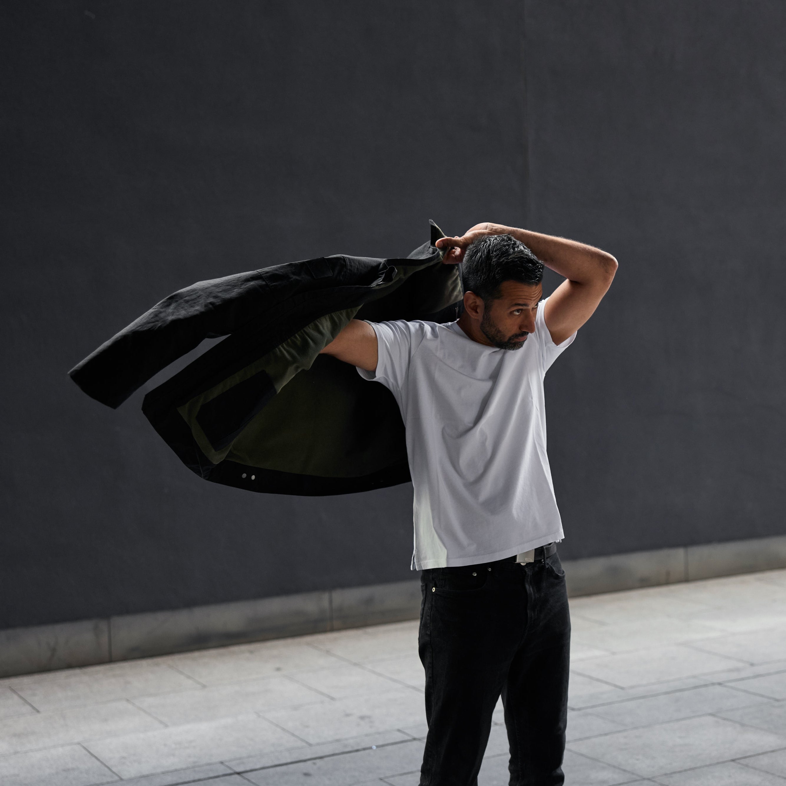 Man putting a black jacket on against a dark wall.