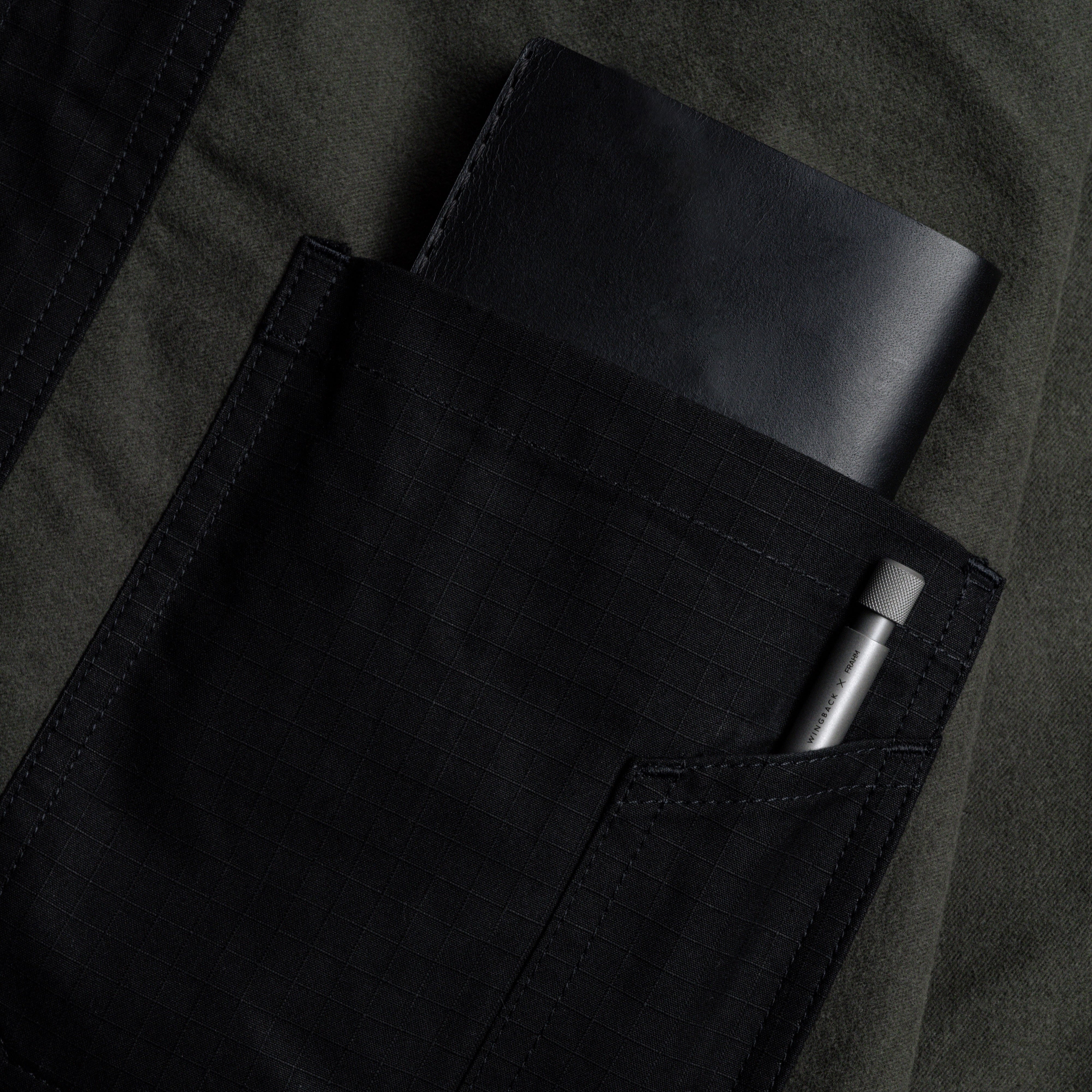 Close-up of a person wearing a dark jacket with a black pocket containing a black notebook and a silver pen.