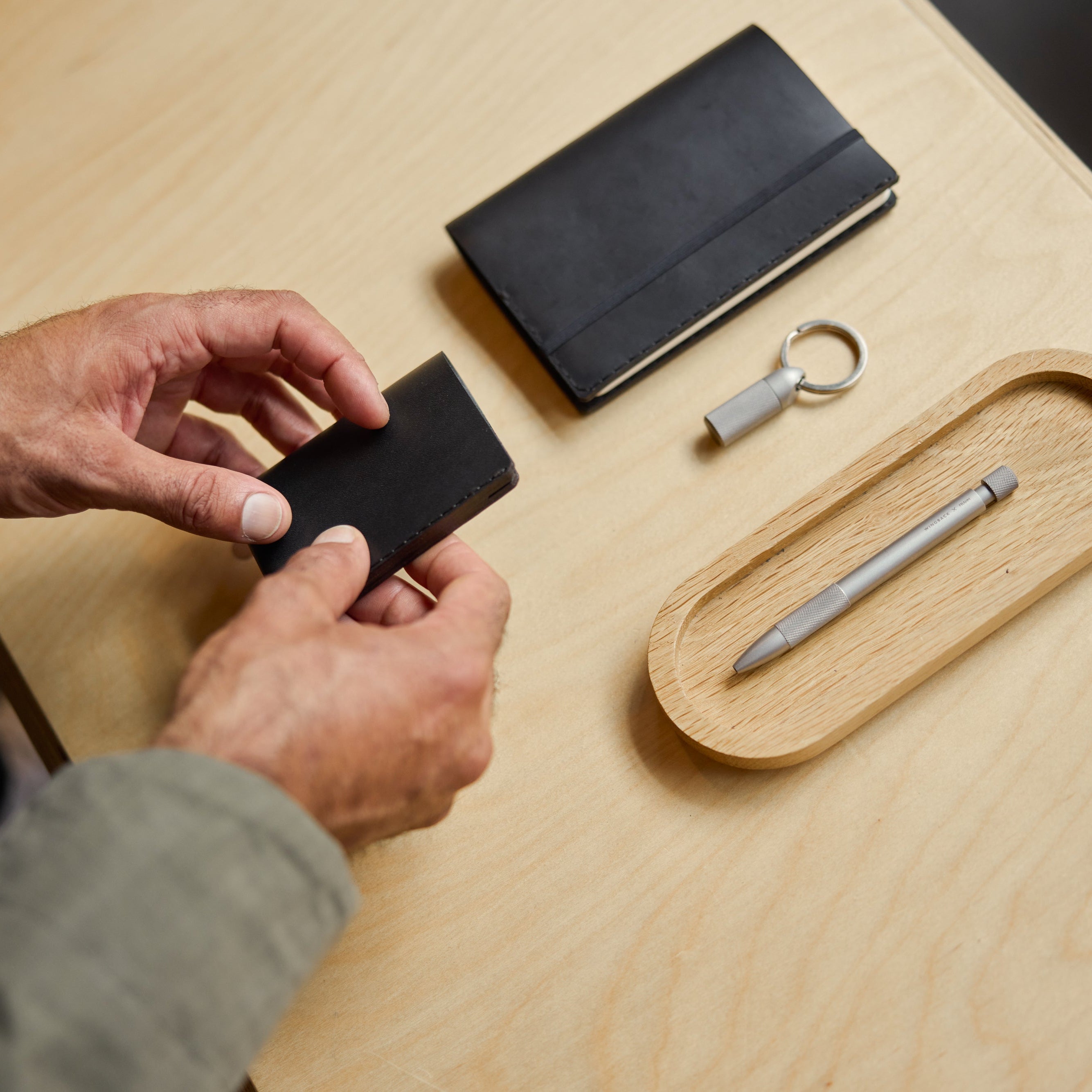 Person holding a black wallet with a wooden tray, pen, and keychain on a light wood surface.