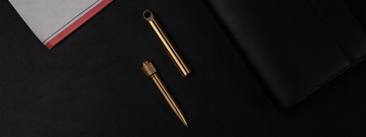 Wingback Pocket Pen in machined brass, cap off, on black leather with an airmail envelope and notebook