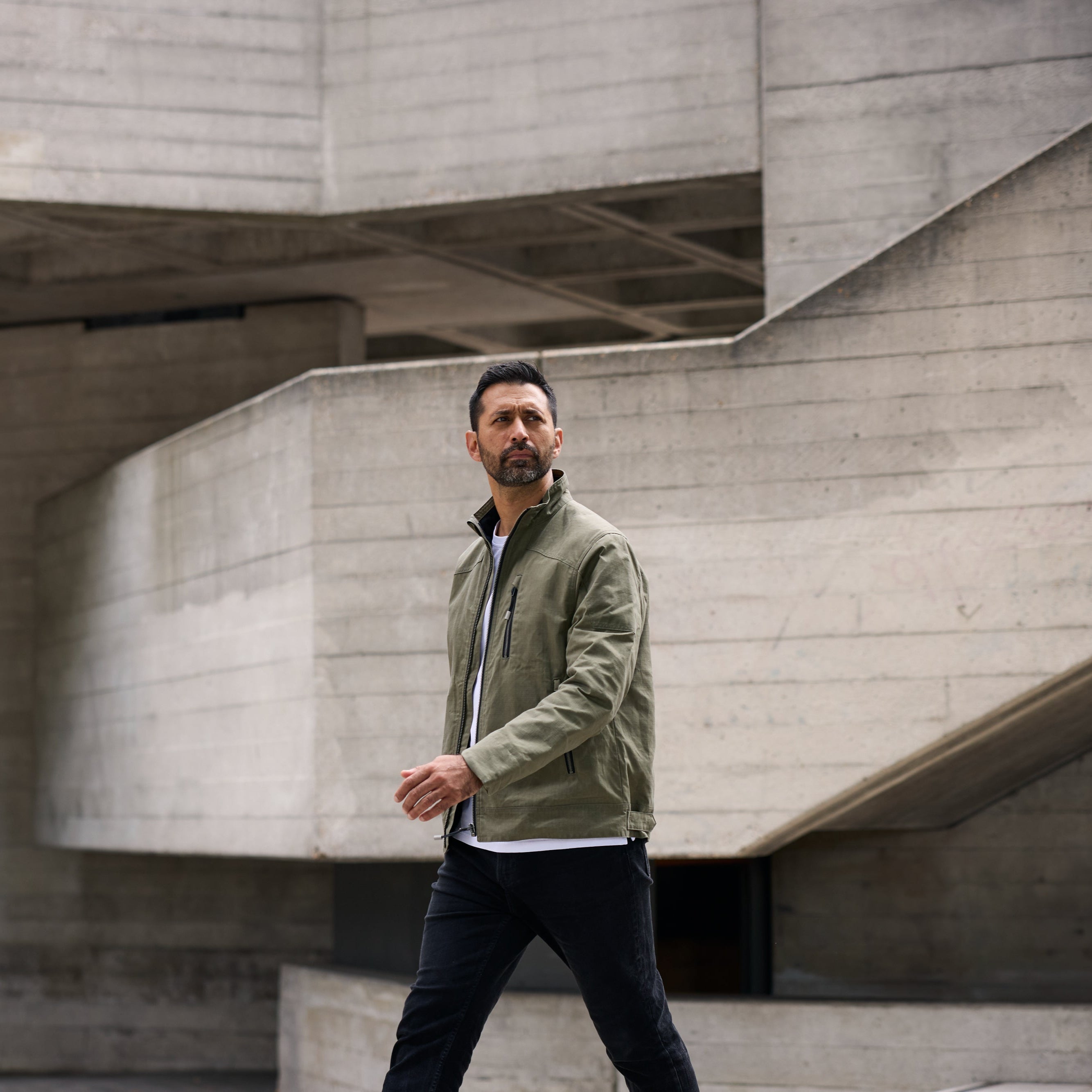 Man wearing a green jacket walking in front of concrete architectural structures.